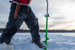 An ice fisher drills a hole on a frozen lake in the low winter sun near Arcora Boutique Hotel & Villas, Muonio.