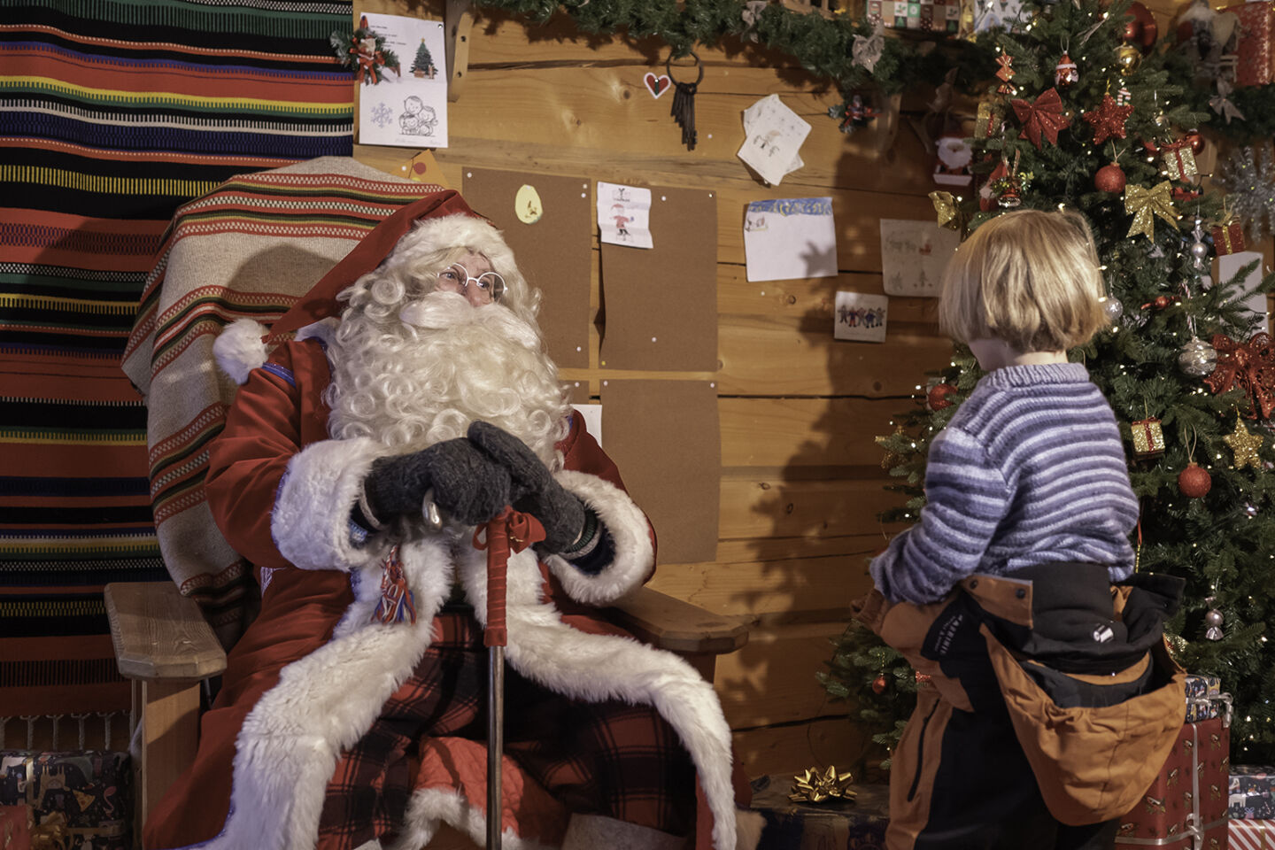 Santa Claus waits in his log cabin as a young guest studies the lit Christmas tree, a festive visit near Arcora, Muonio.