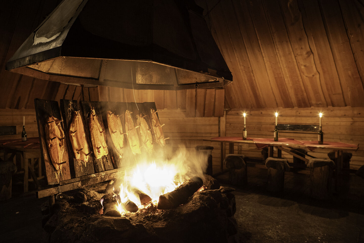 Salmon roasts on wooden planks by an open fire in a log kota, candlelit tables set for dinner in the wilderness near Arcora.