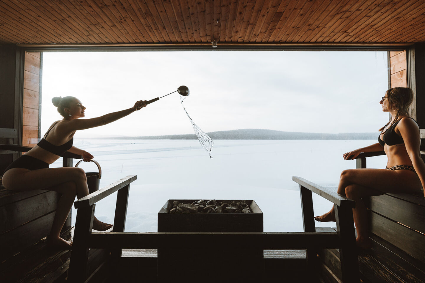 A guest throws water on the stones in a panorama sauna above a frozen lake at Arctic Sauna World, Muonio, Finnish Lapland.